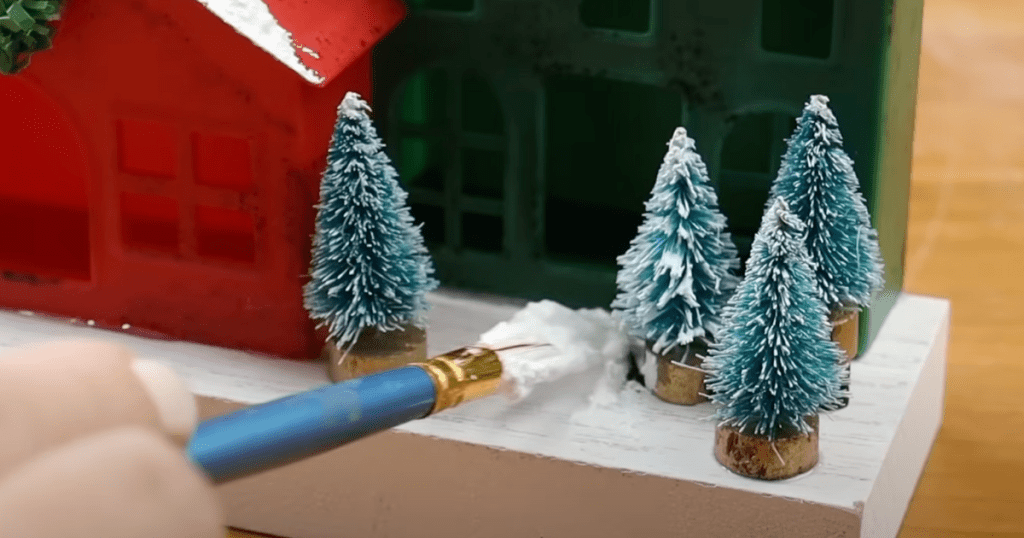 using a paint brush to add decoart snow tex onto a base that has little bottle brush trees glued to it from dollar tree