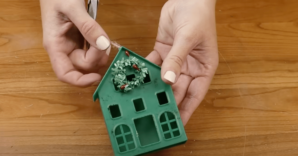 a woman with painted finger nails holding a small plastic green house ornament with a tiny wreath on it from dollar tree on a wood background